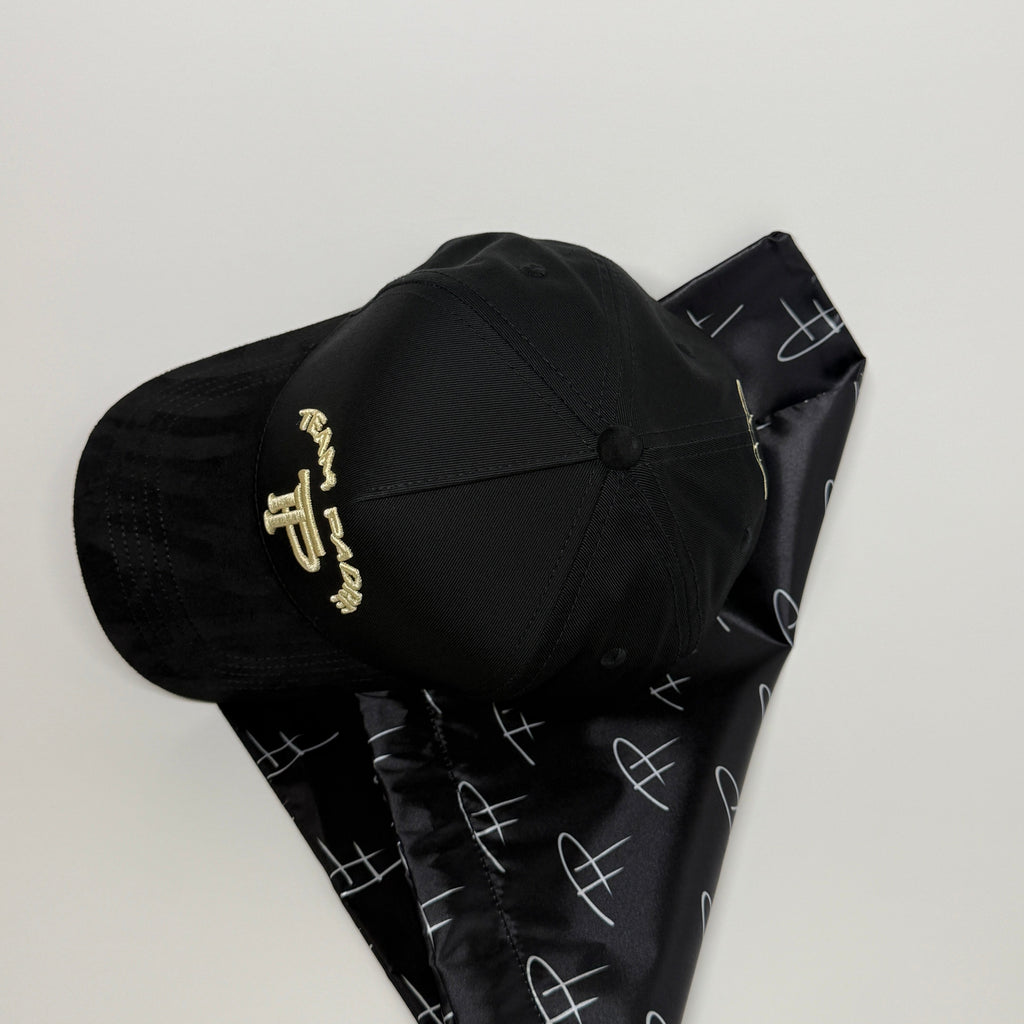 A black baseball cap with gold embroidered lettering, resting on a black neck gaiter with a white abstract pattern, set against a plain light gray background.