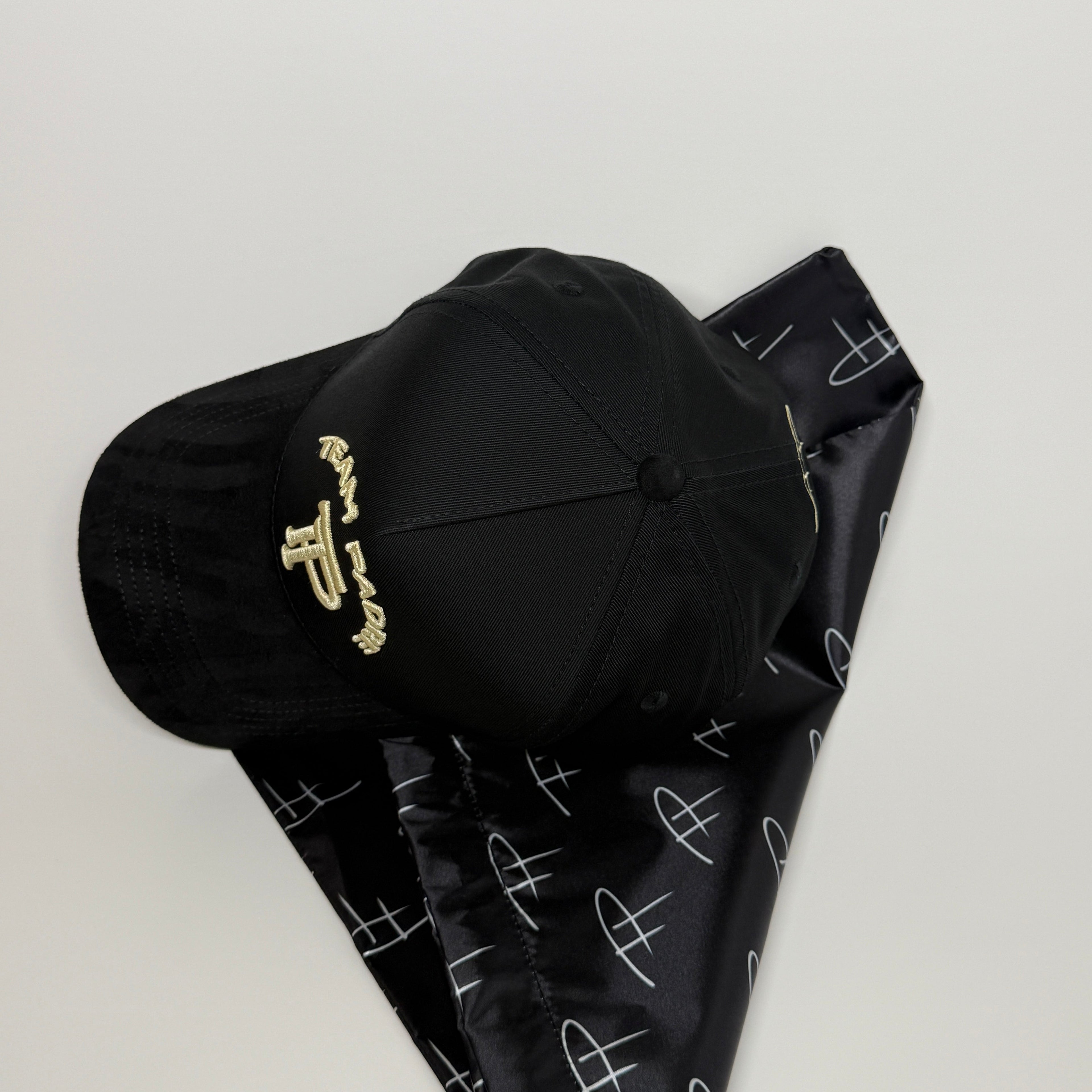 A black baseball cap with gold embroidered lettering, resting on a black neck gaiter with a white abstract pattern, set against a plain light gray background.